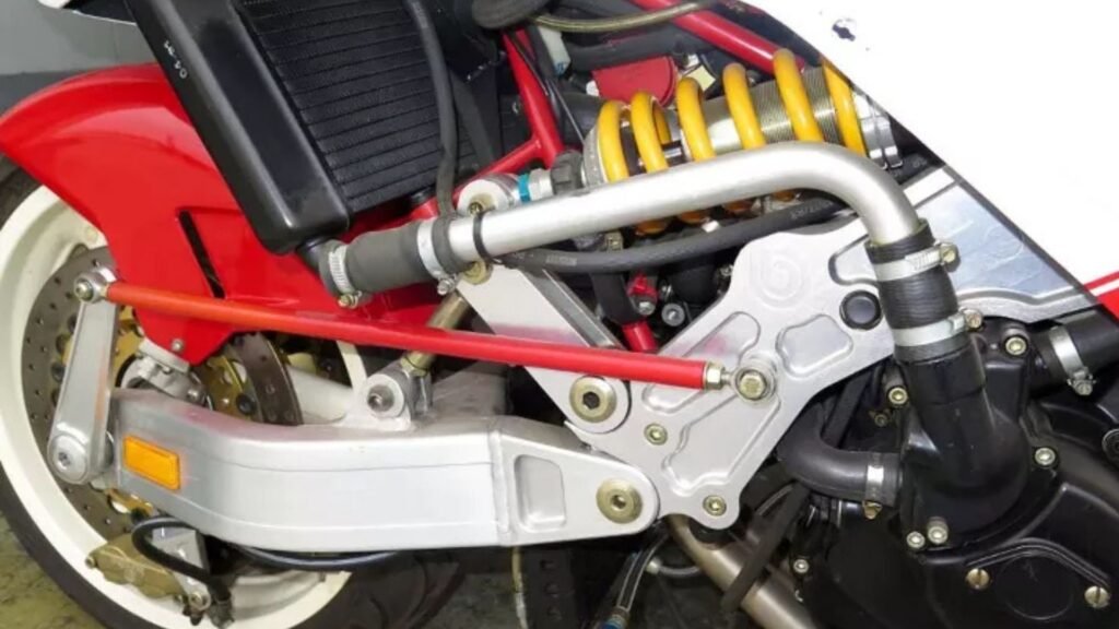 O braço duplo da suspensão é ligado ao quadro e seu amortecimento é feito por um robusto amortecedor. O braço vermelho é o responsável por esterçar a motocicleta.